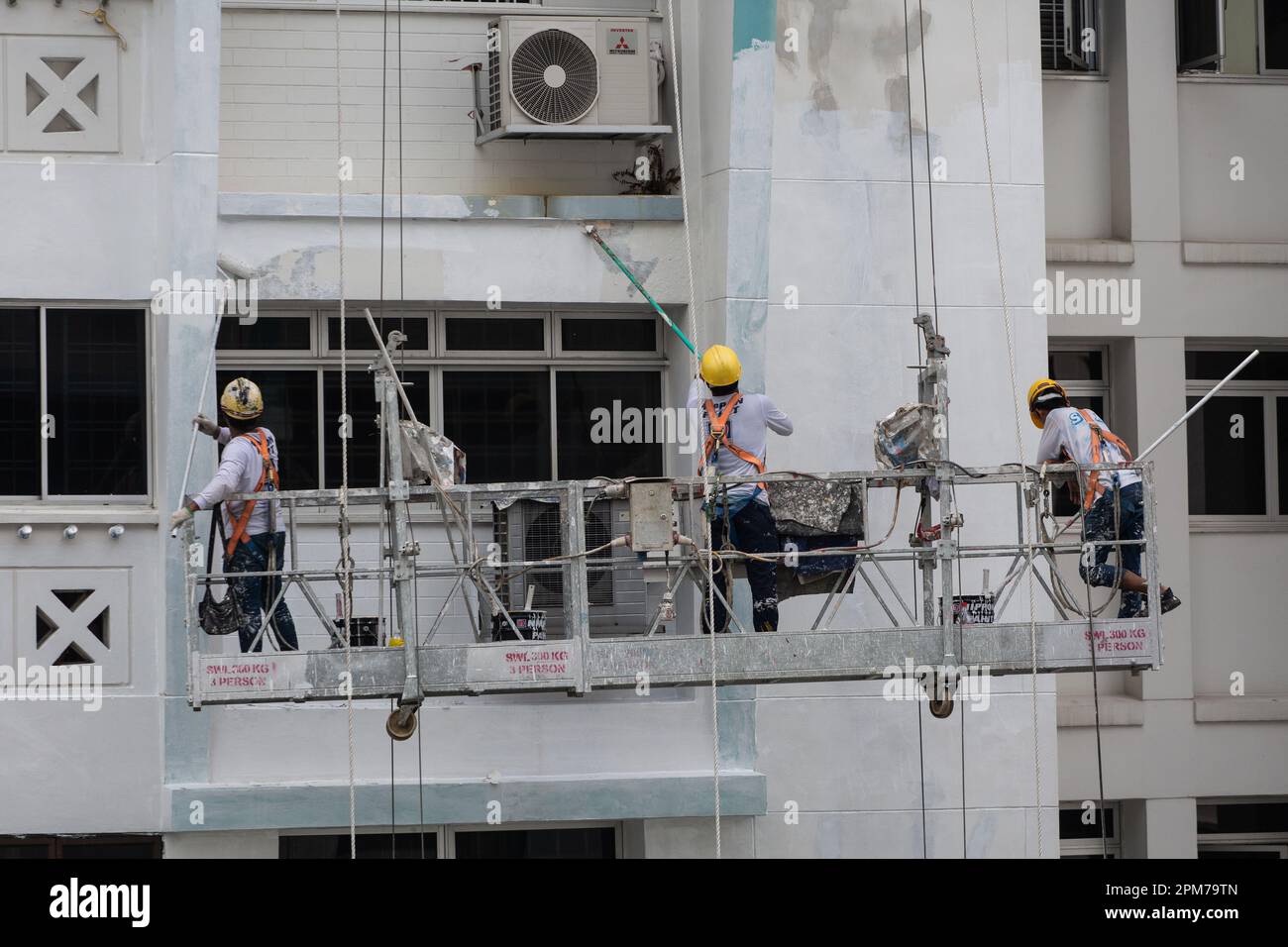 Drei Arbeiter auf den Gerüstaufzügen in der Höhe, die ihr Bestes tun, um Schälfarbe abzukratzen und neue Farbe auf der Fassade des Gebäudes zu beschichten. Stockfoto