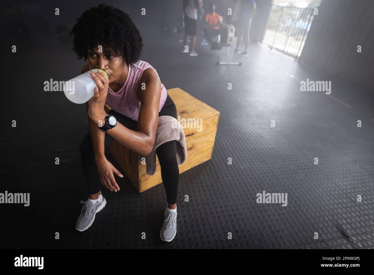 Blick aus dem hohen Winkel auf müde, birassische junge Frau, die Wasser trinkt, während sie im Fitnessstudio auf einer Holzkiste sitzt Stockfoto