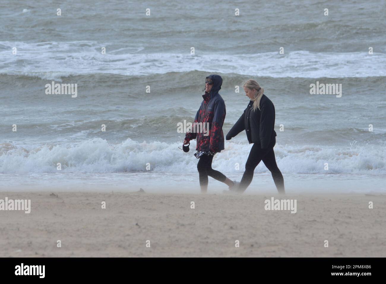 Storm Noa, Boscombe, Bournemouth, Dorset, Vereinigtes Königreich, 12. April 2023, Wetter. Zwei junge Frauen, die morgens am Strand spazieren, haben starke Winde und stürmische Bedingungen am Meer. Kredit: Paul Biggins/Alamy Live News Stockfoto