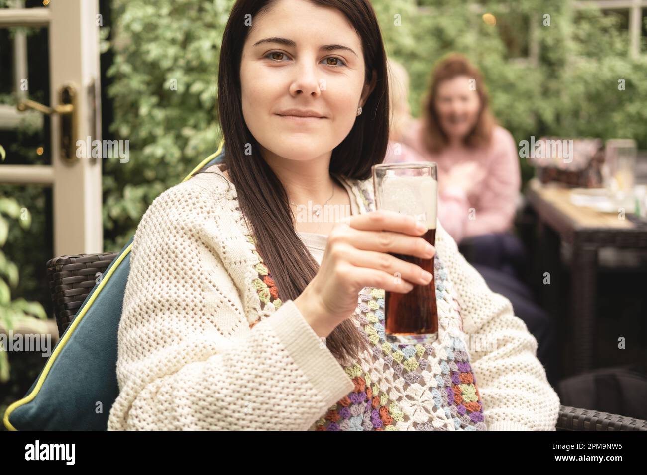 Porträt einer wunderschönen brünetten Frau und einer neuen Mutter, die in die Kamera schaut und nach einem Spaziergang mit ihrer Familie im Biergarten im Pub ein alkoholfreies Getränk trinkt Stockfoto