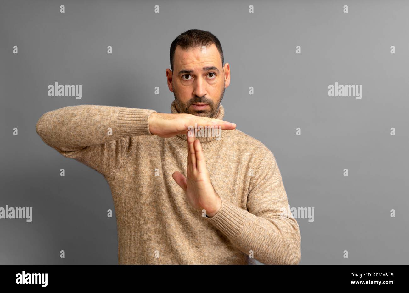 Bärtiger hispanischer Mann in seinen 40s Jahren mit beigefarbenem Rollkragenpullover, der die Auszeit-Geste machte, während er ernsthaft in die Kamera schaute, isoliert im grauen Studio Stockfoto