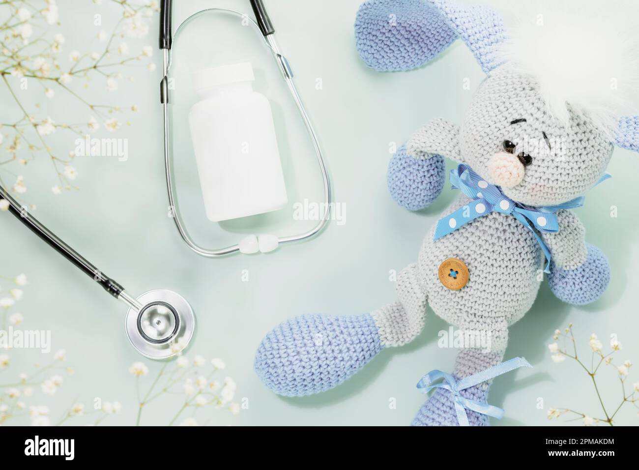 Kindergesundheitsfürsorge. Beilagen für Kinder, Vitamine oder Pillen in weißer Flasche mit Stethoskop und niedlichem gestricktem Spielzeughasen auf blauem Hintergrund mit weißen Blumen Stockfoto
