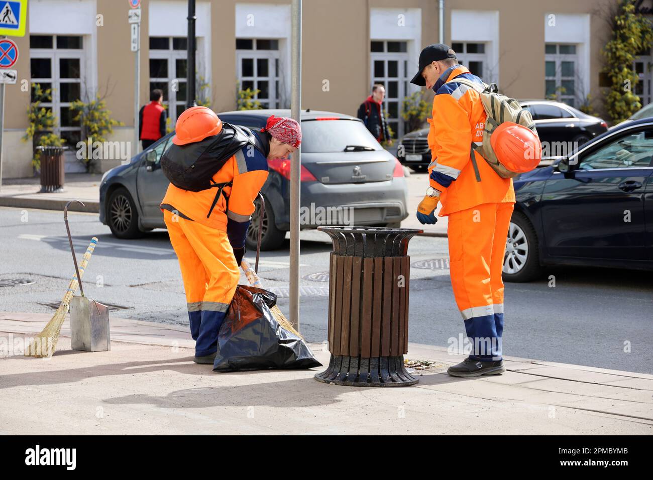 Zwei Hausmeister in orangefarbener Uniform durchkämmen die Straße auf dem Hintergrund von Autos. Mann und Frau, die die Frühjahrsstadt säubern Stockfoto
