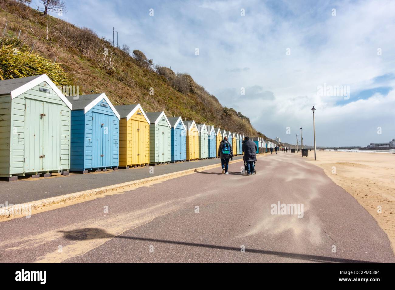 Die Menschen gehen einen Pfad vor den Strandhütten am Bournemouth Beach in Dorset, Großbritannien, unter stürmischem, grauem Himmel mit Sand, der im Wind weht Stockfoto