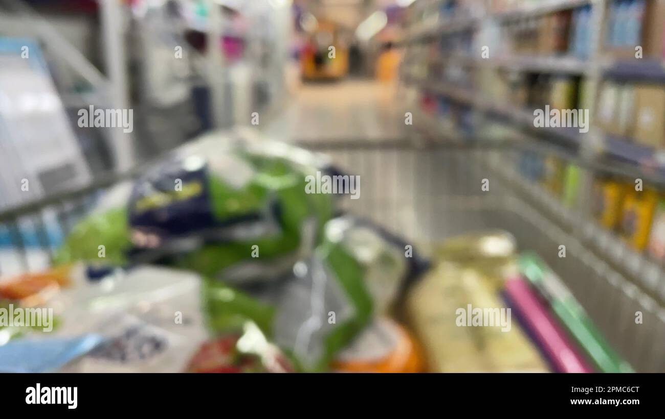 Ein Einkaufswagen in einem Supermarkt aus einem Warenkorb, der unscharf ist, mit unbekannten Leuten, die Produkte in einem Supermarkt-Regal wählen. Essen kaufen und Stockfoto