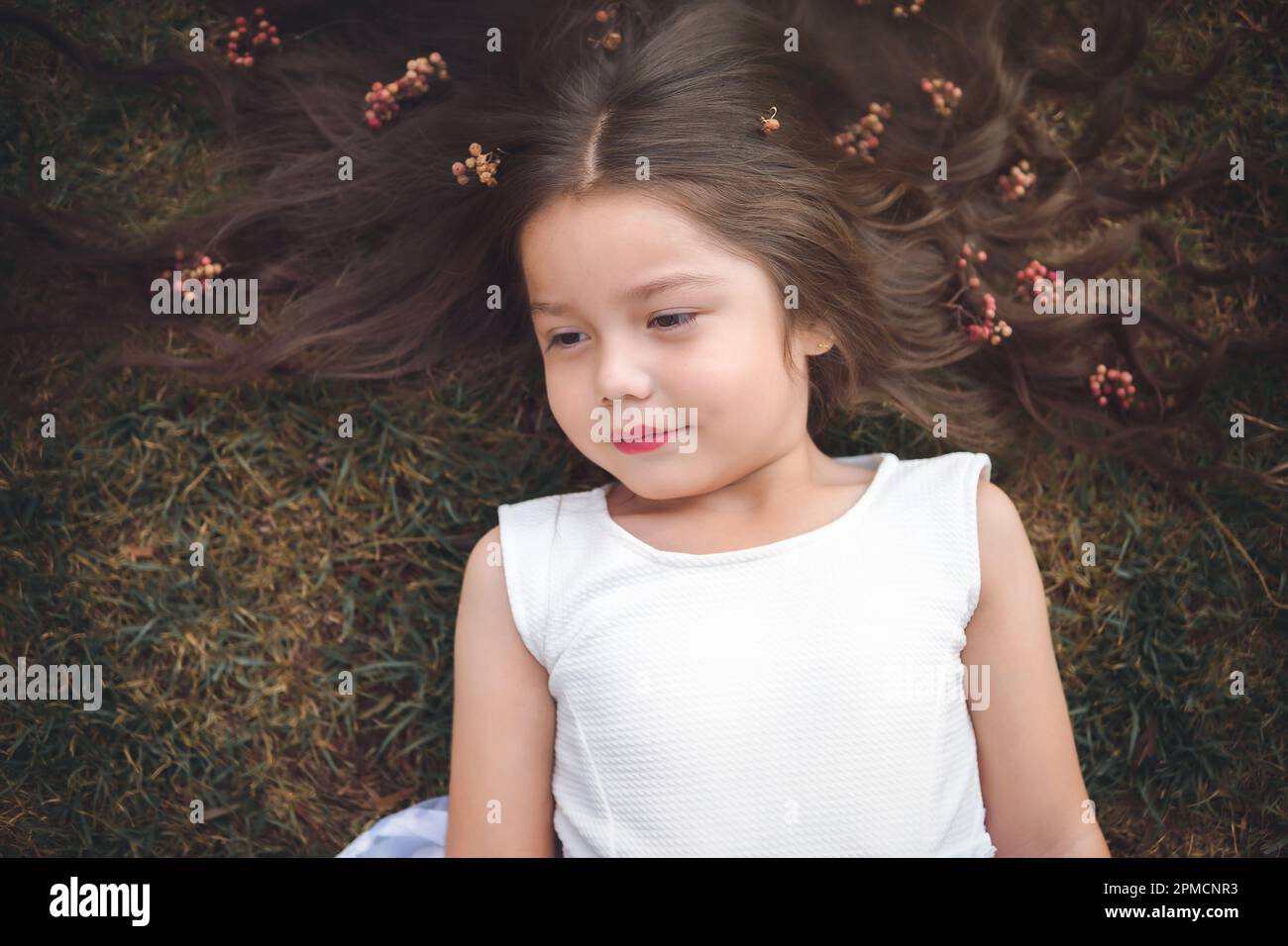 Ein kleines Mädchen in weißem Kleid, das auf dem Gras liegt, lächelt und ihr Haar ist sehr lang und ein Prinzessinnen-Motto für den Kindertag. Stockfoto