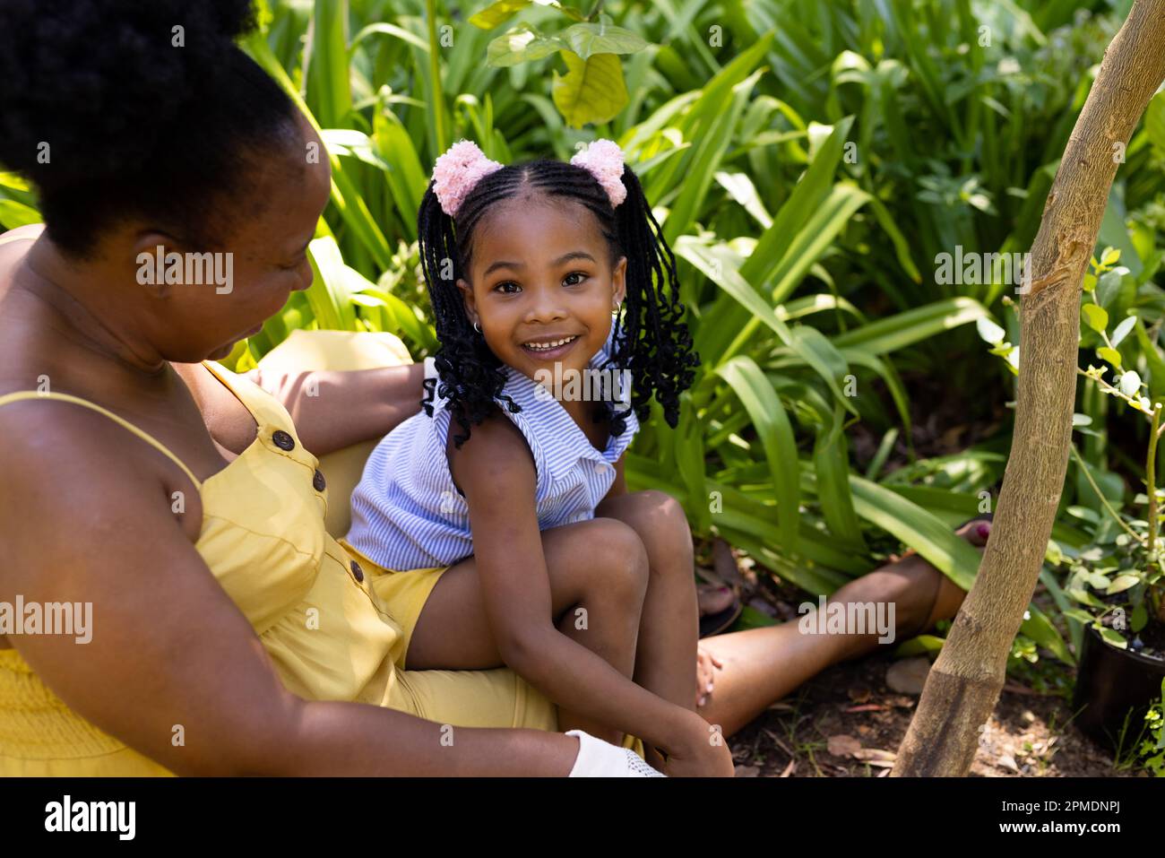 afroamerikanisches lächelndes Mädchen, das mit Großmutter bei Pflanzen sitzt, während es im Garten Gartenarbeit macht Stockfoto