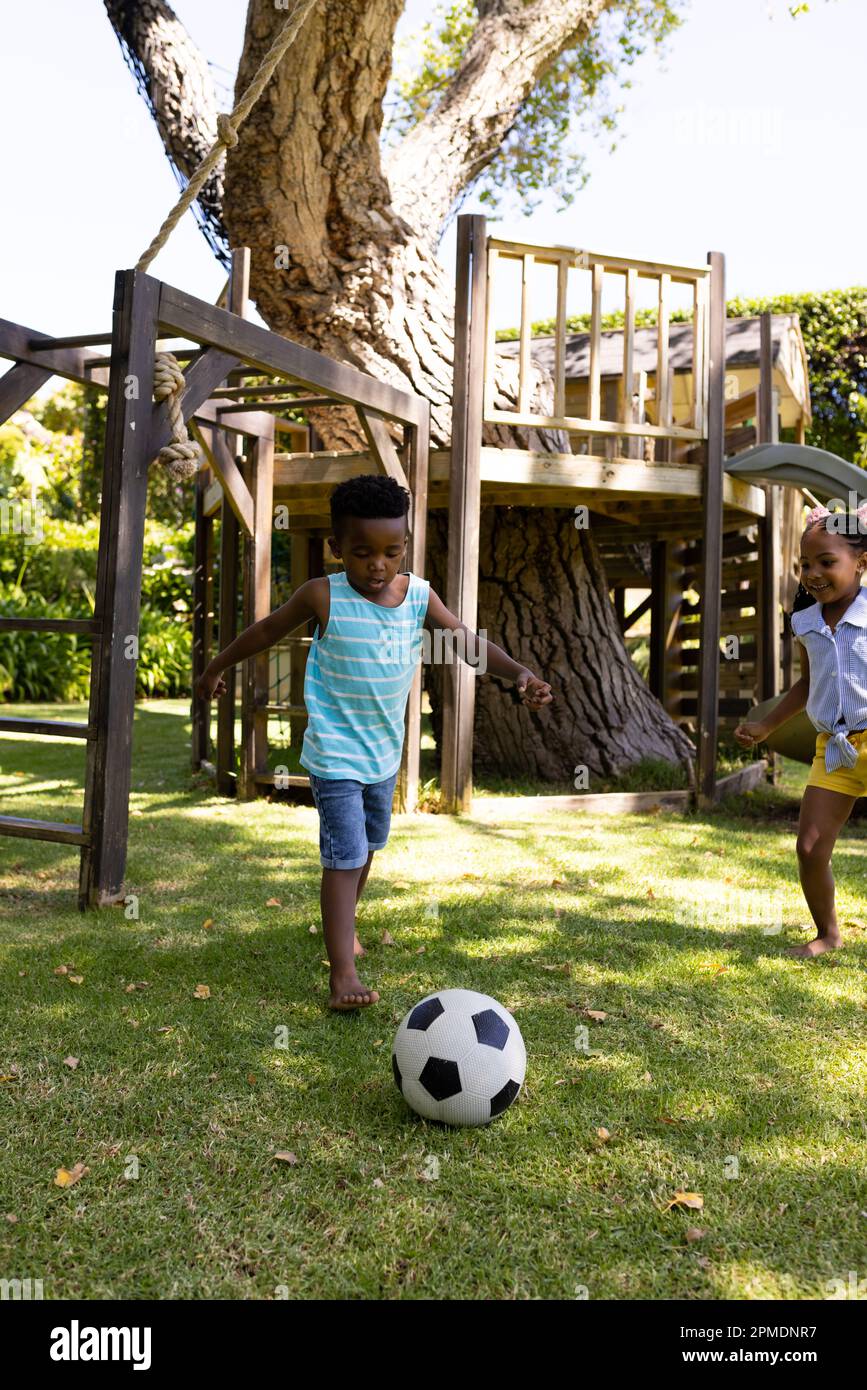 Fröhliche afroamerikanische Geschwister, die auf dem grasbedeckten Feld im Park Fußball spielen Stockfoto
