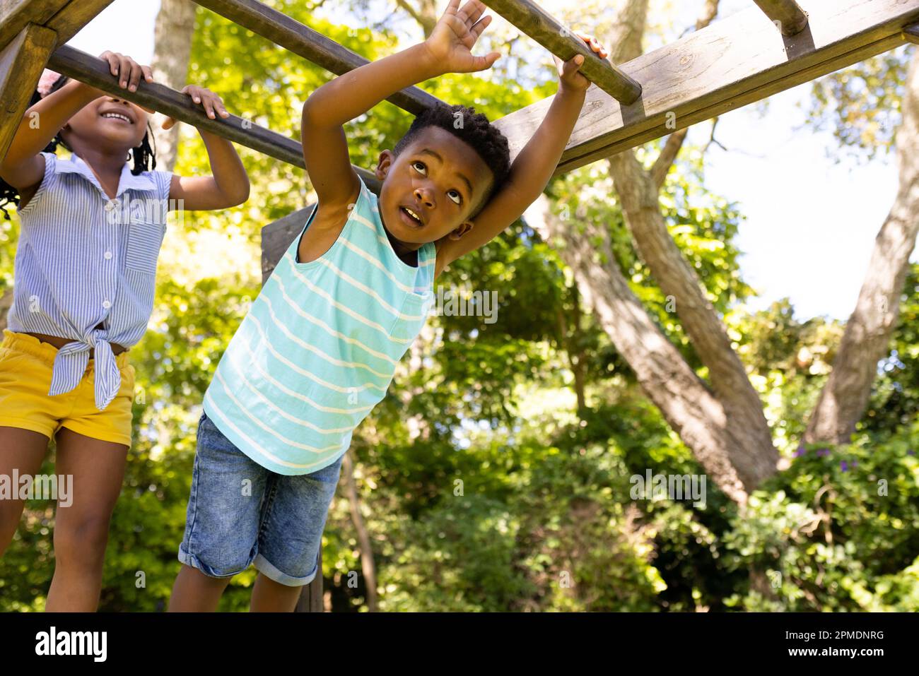 Blick aus flachem Winkel auf die verspielten afroamerikanischen Geschwister, die an den Affenbars auf dem Spielplatz hängen Stockfoto