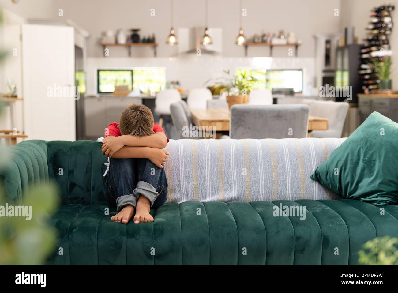 Ein weißer Junge in voller Länge umarmt die Knie, während er auf dem Sofa im Wohnzimmer sitzt, Kopierbereich Stockfoto