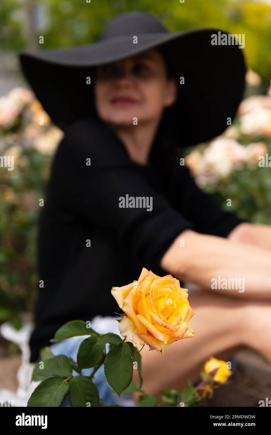Verschwommenes Bild einer sitzenden Frau mit Hut und Shorts, die wegschaut, neben gelben Rosen, selektiver Fokus. Stockfoto