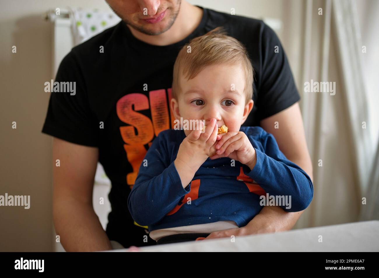 Ein kleines Kleinkind, das von seinem Vater gehalten wird und ein Stück Brot isst Stockfoto