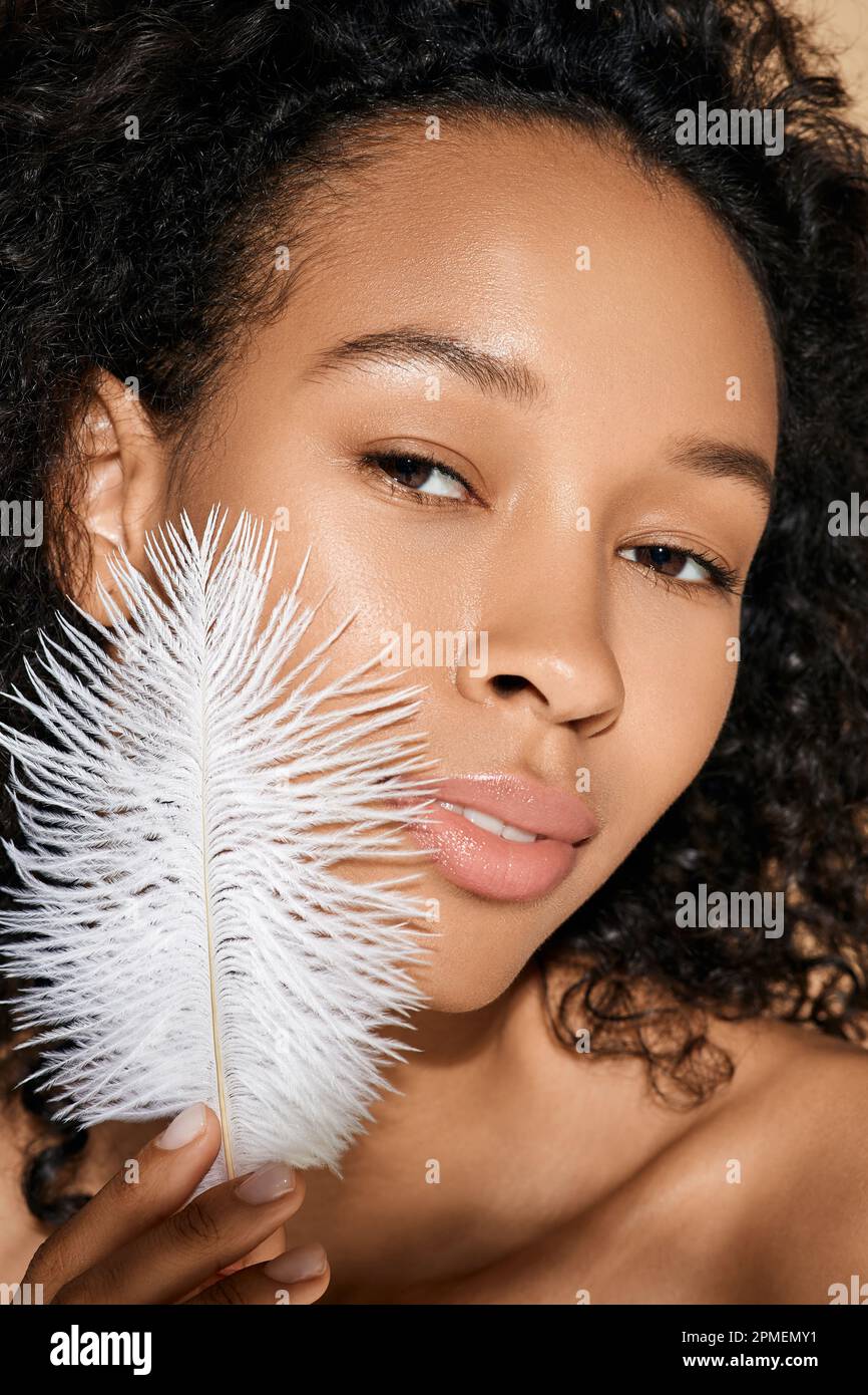 Gesichtshaut-Pflege. Die afroamerikanische Frau zeigt glatte und weiche Haut, indem sie ihr Gesicht mit weißer Feder berührt Stockfoto