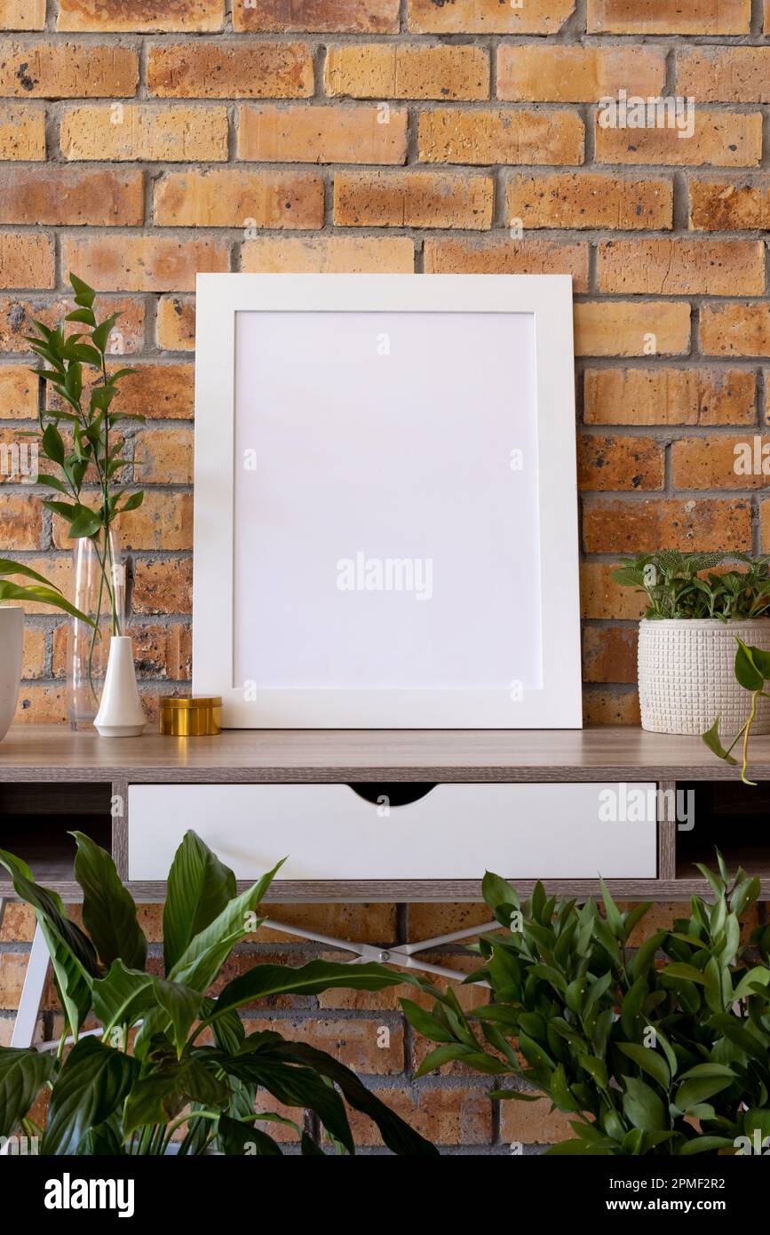 Vertikaler, leerer weißer Holzrahmen mit Kopierraum und Pflanzen in Töpfen auf dem Schreibtisch an der Ziegelwand Stockfoto