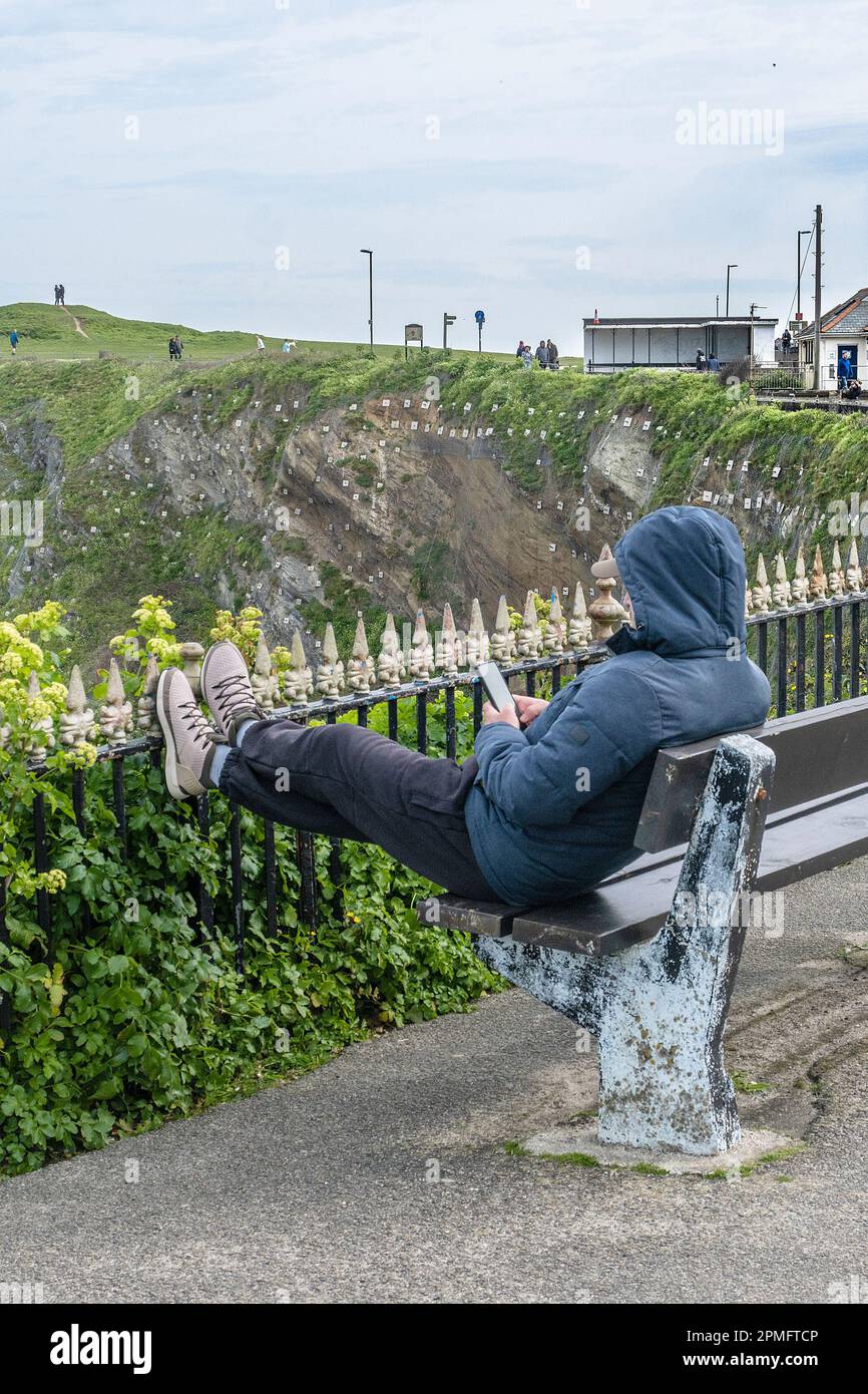 Ein Mann, der sich mit den Füßen auf Geländern entspannt und mit seinem Mobiltelefon in Newquay in Cornwall, Großbritannien, auf einer Bank sitzt. Stockfoto