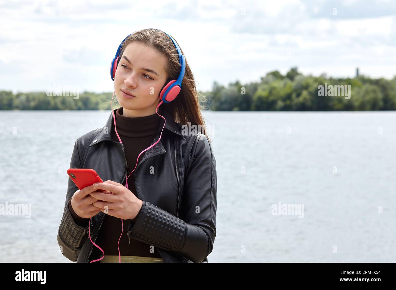 Die junge kaukasische Frau mit blauroten Kopfhörern hört auf dem Smartphone am Flussufer Musik und Tippen. Portrait Foto von glücklichen Hipster Mädchen rel Stockfoto