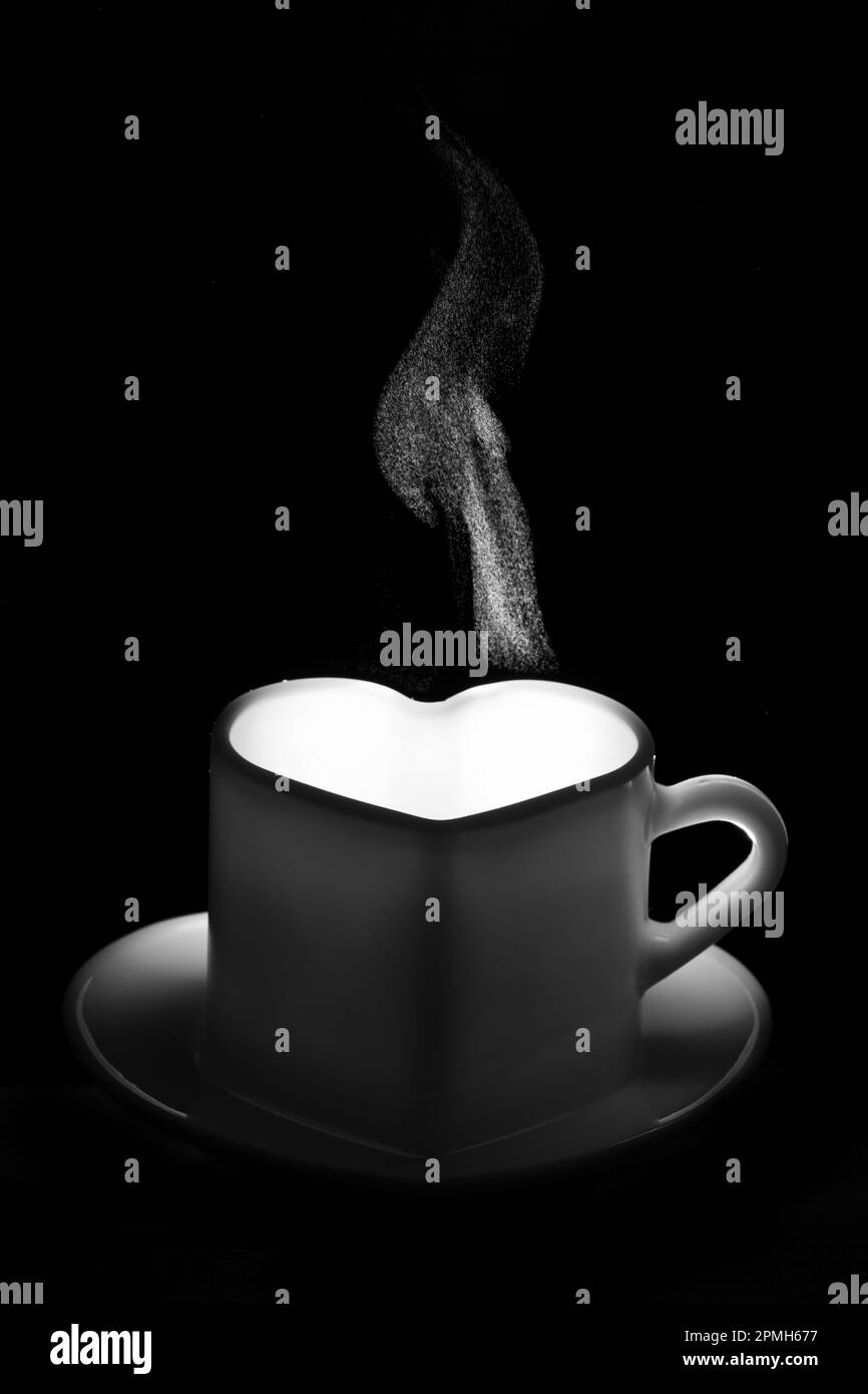 Ein weißer herzförmiger Becher mit einem warmen Getränk und ausgefallenem lockigem Dampf. Eine dampfende Kaffeetasse auf schwarzem Hintergrund. Morgenkaffee. Kaffee für Stockfoto