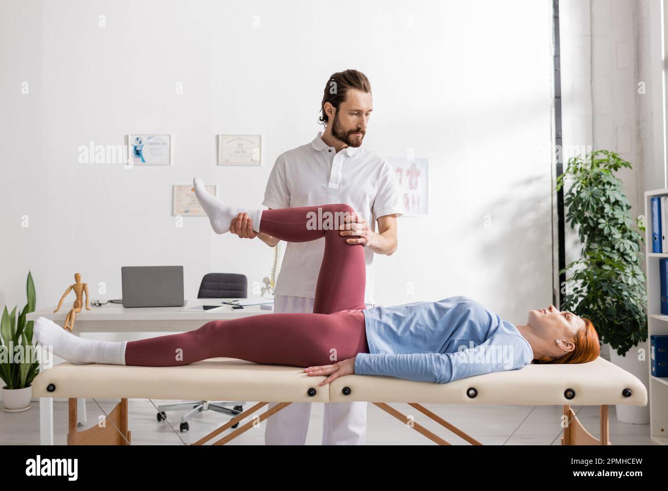 Rehabilitologe, der schmerzhafte Beine einer Frau beugt, die auf dem Massagetisch im Sprechzimmer liegt, Stockbild Stockfoto