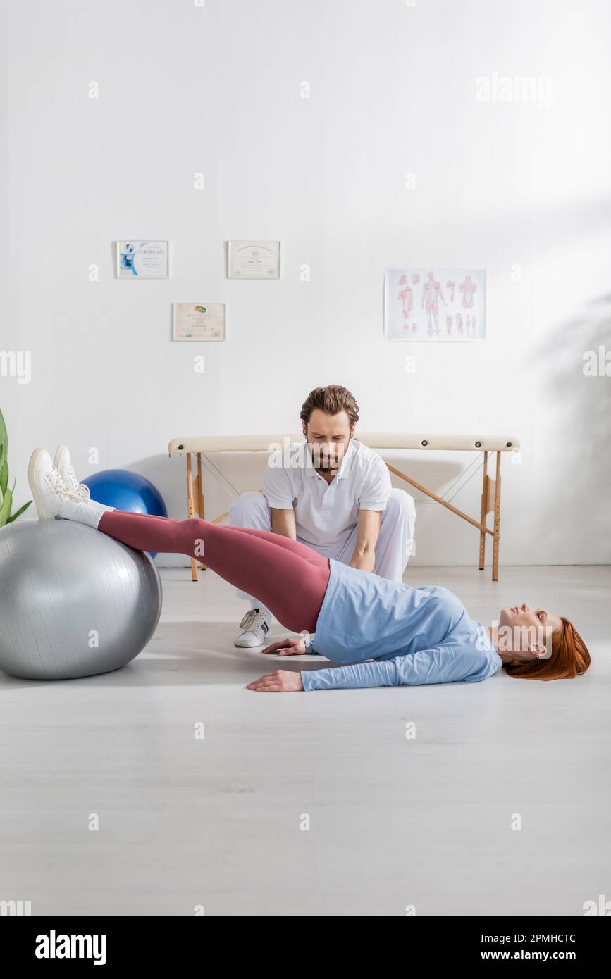 Bärtige Physiotherapeutin in der Nähe einer Frau, die auf dem Boden liegt und mit einem Fitnessball in der Reha trainiert, Stockbild Stockfoto