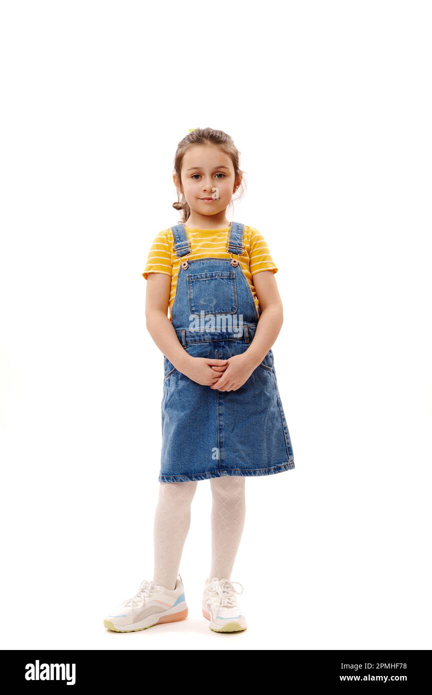 Ein langes Porträt eines hübschen kleinen Mädchens in Jeansbekleidung, das isoliert auf weißen Hintergrund in die Kamera schaut Stockfoto
