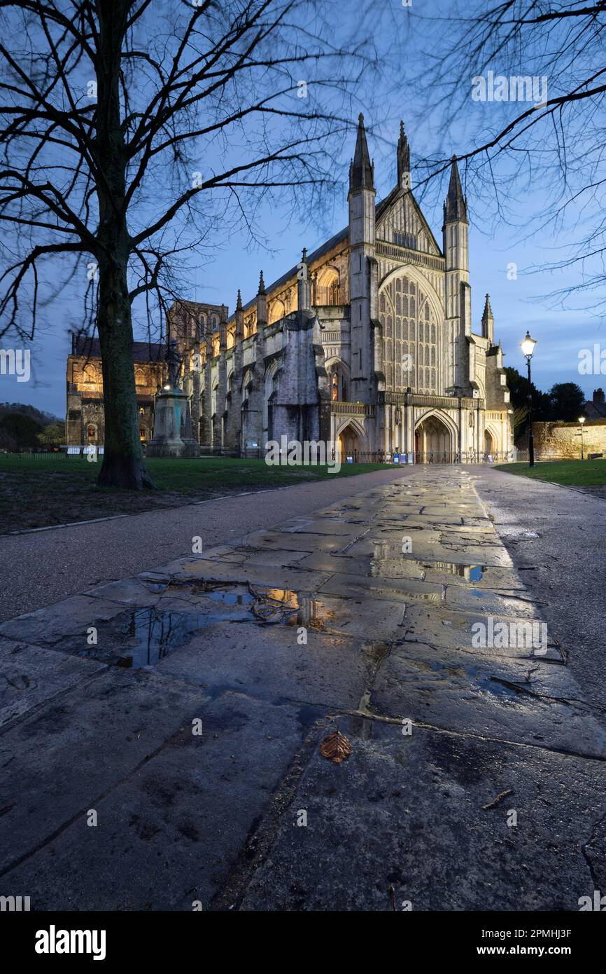 Die Kathedrale von Winchester erstrahlte nachts im Winter mit Reflexionen auf nassem Bürgersteig im Vordergrund, Winchester, Hampshire, England, Großbritannien Stockfoto
