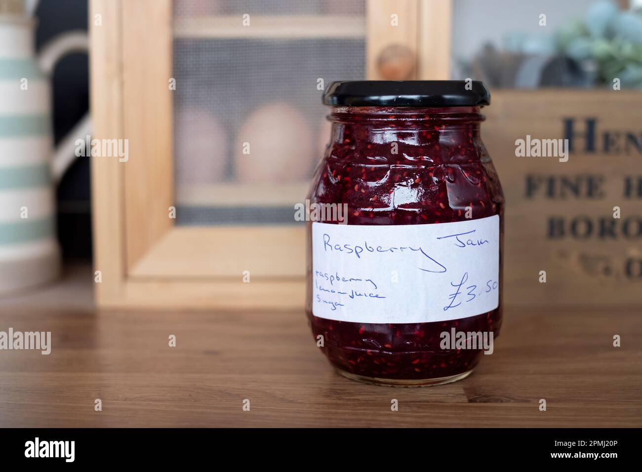 Ein Glas hausgemachter Himbeermarmelade. Der Mixbecher verfügt über ein handgeschriebenes Etikett, auf dem die Zutaten und der Preis der Marmelade aufgeführt sind. Stockfoto