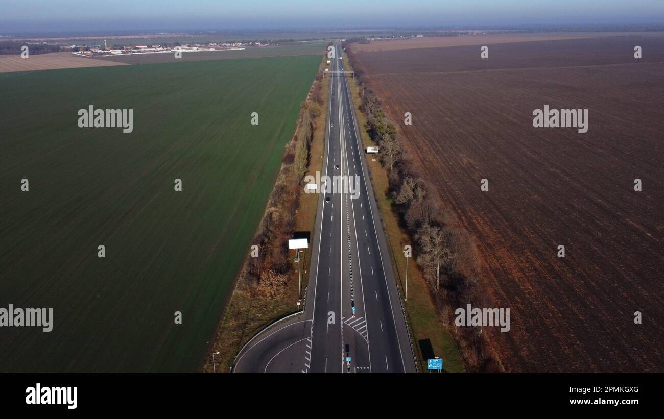 Wunderschöne Panoramalandschaft. Schwarz gepflasterte Autobahn mit weißen Markierungen, viele vorbeifahrende Autos, große landwirtschaftliche Felder mit grüner landwirtschaftlicher Ernte und gepflügerter schwarzer Boden an einem sonnigen Herbsttag. Stockfoto