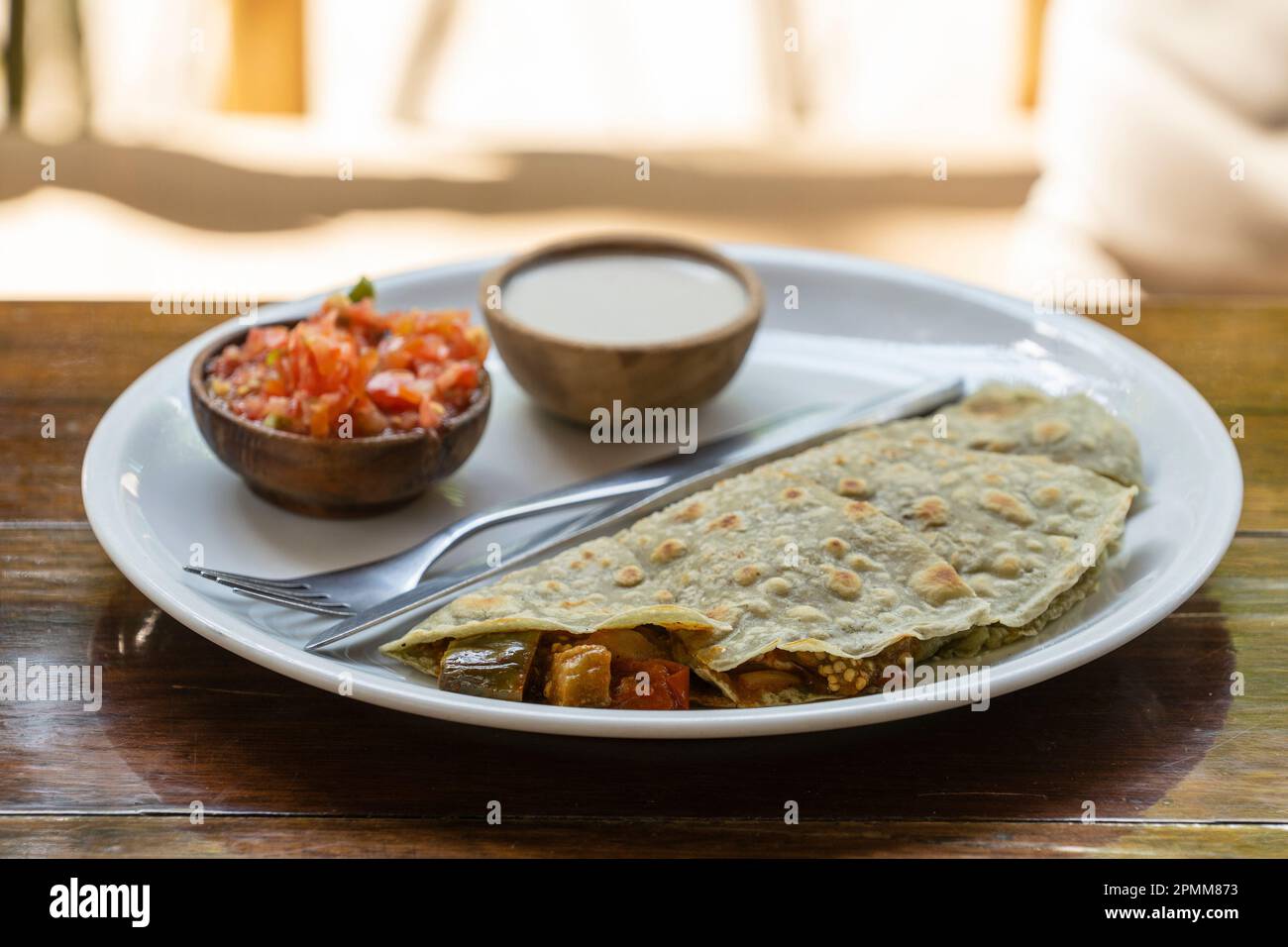 Quesadillas aus Spiruline Tortilla mit Cashewkäse, Auberginen, Tomaten und Sauerrahm und Tomatensalsa auf einem Holztisch im tropischen Café in Thailand, Stockfoto