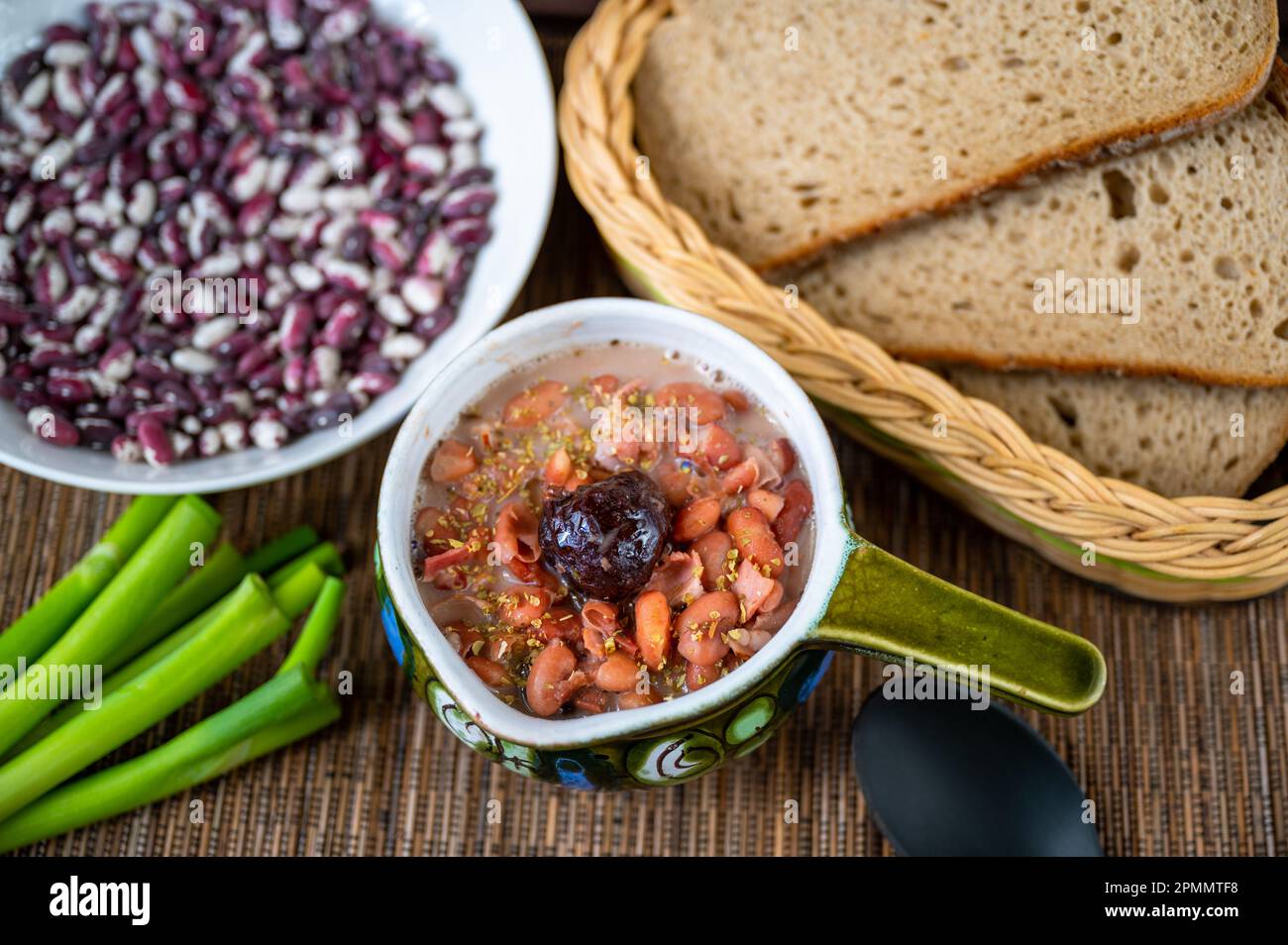 Suppe aus Bohnen und Pflaumen in der Schüssel, rohe Bohnen auf dem Teller, grüne Zwiebelsprossen, Brot im Korb, rustikaler Hintergrund. Stockfoto