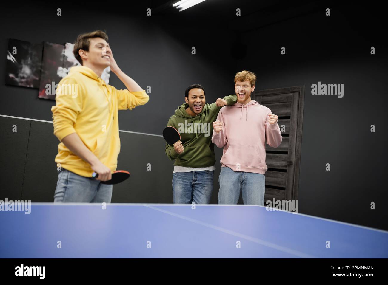Aufgeregte, gemischtrassige Männer stehen neben einem verschwommenen Freund mit Tennisschläger im Spielclub, Aktienbild Stockfoto