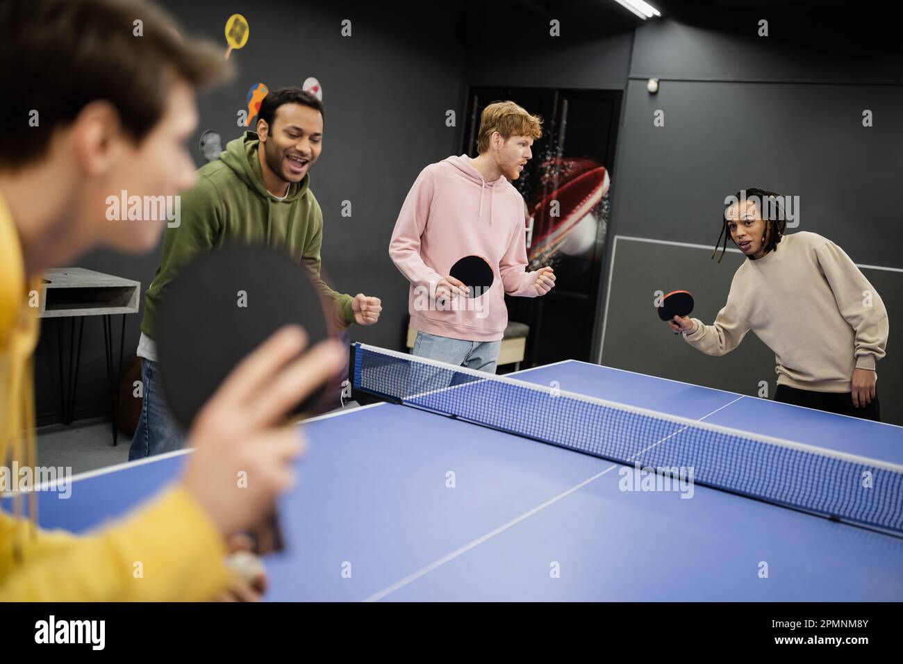 Aufgeregte Männer, die in der Nähe von Freunden stehen und Tischtennis spielen, im Spielclub, Aktienbild Stockfoto