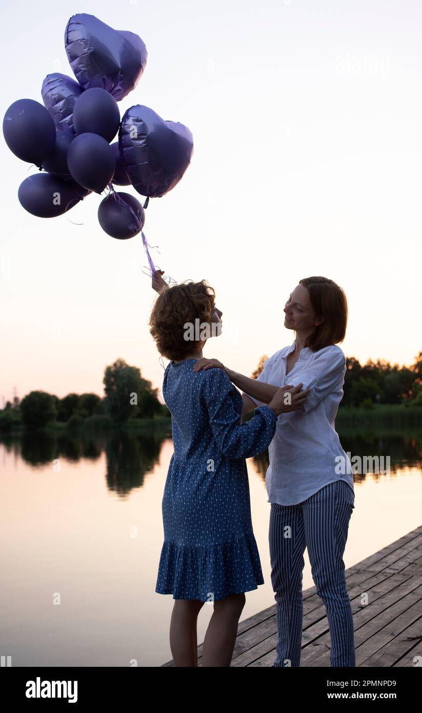 mutter umarmt ihre Erwachsene Tochter - einen Teenager, der einen Haufen Ballons in der Hand hält und bei Sonnenuntergang am See steht. Zwei Generationen unter guten Bedingungen. Stockfoto