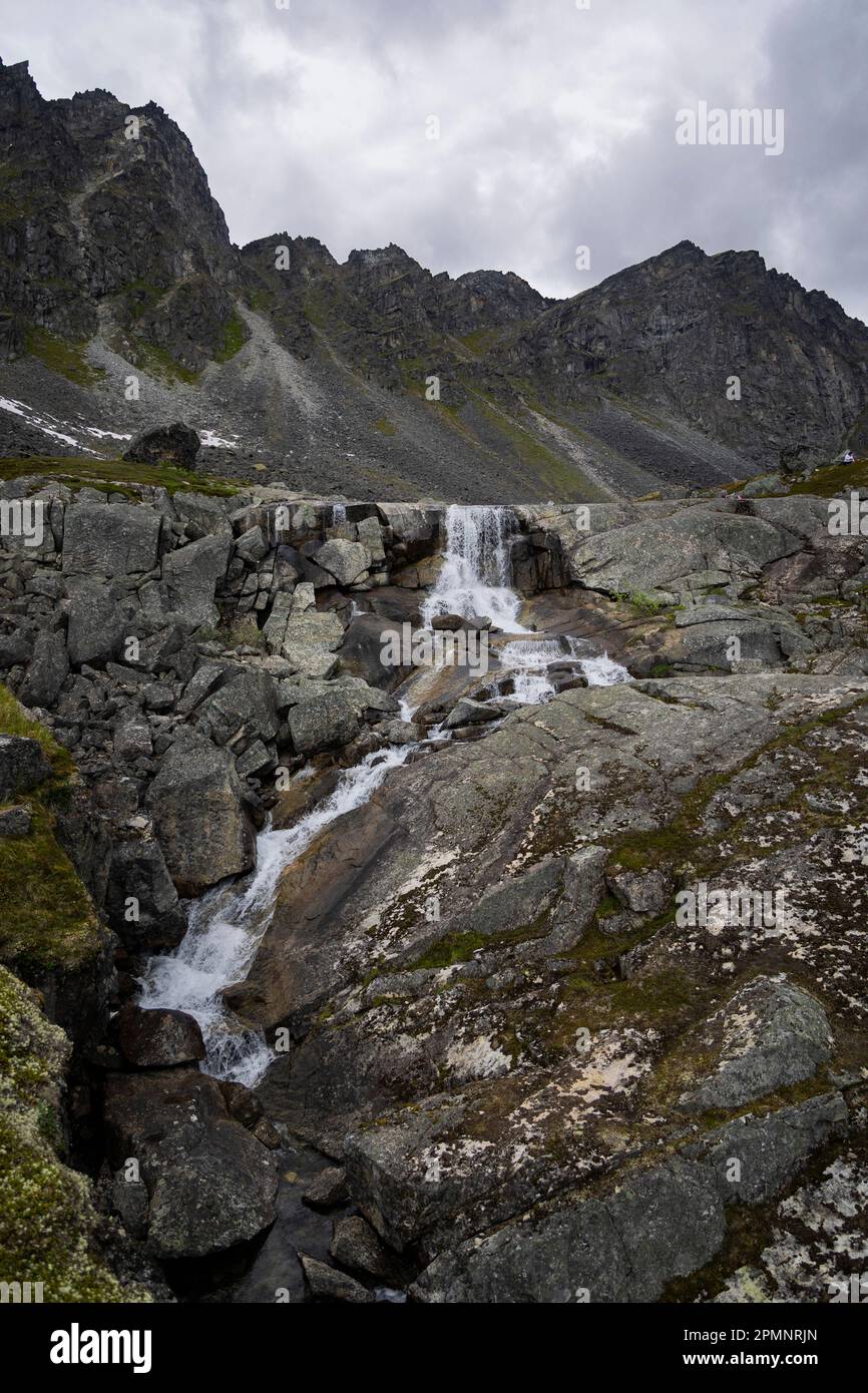 Gletscherwasserfall über felsigem Gelände am Archangel Hatcher Pass mit Granitgipfeln im Hintergrund unter einem grauen, wolkigen Himmel bei Inde... Stockfoto