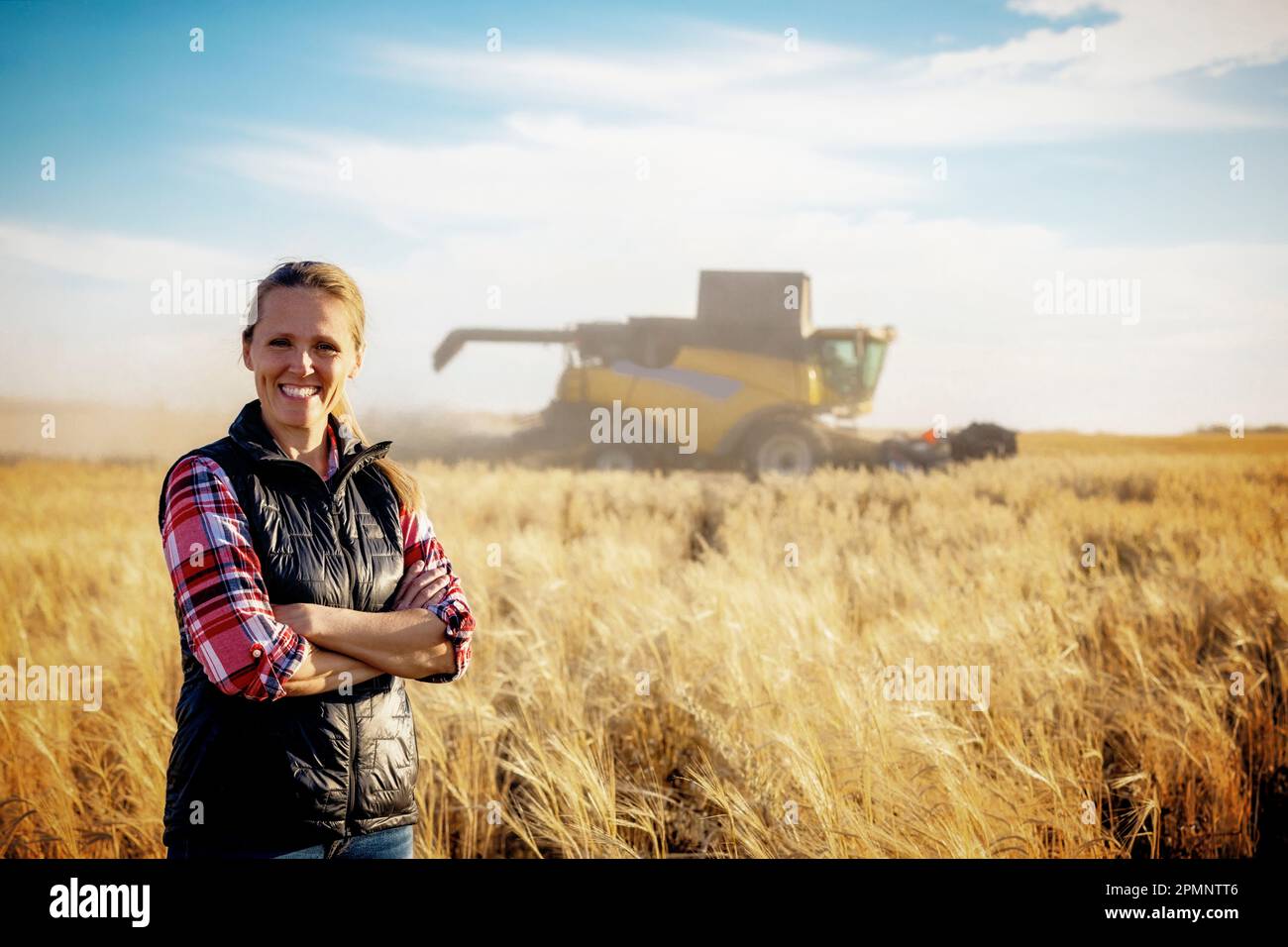 Porträt einer Reifen Landwirtschaftsfrau, die auf einem Getreidefeld steht und während der Ernte für die Kamera posiert, während ein Mähdrescher bei Sonnenuntergang im Hintergrund arbeitet Stockfoto