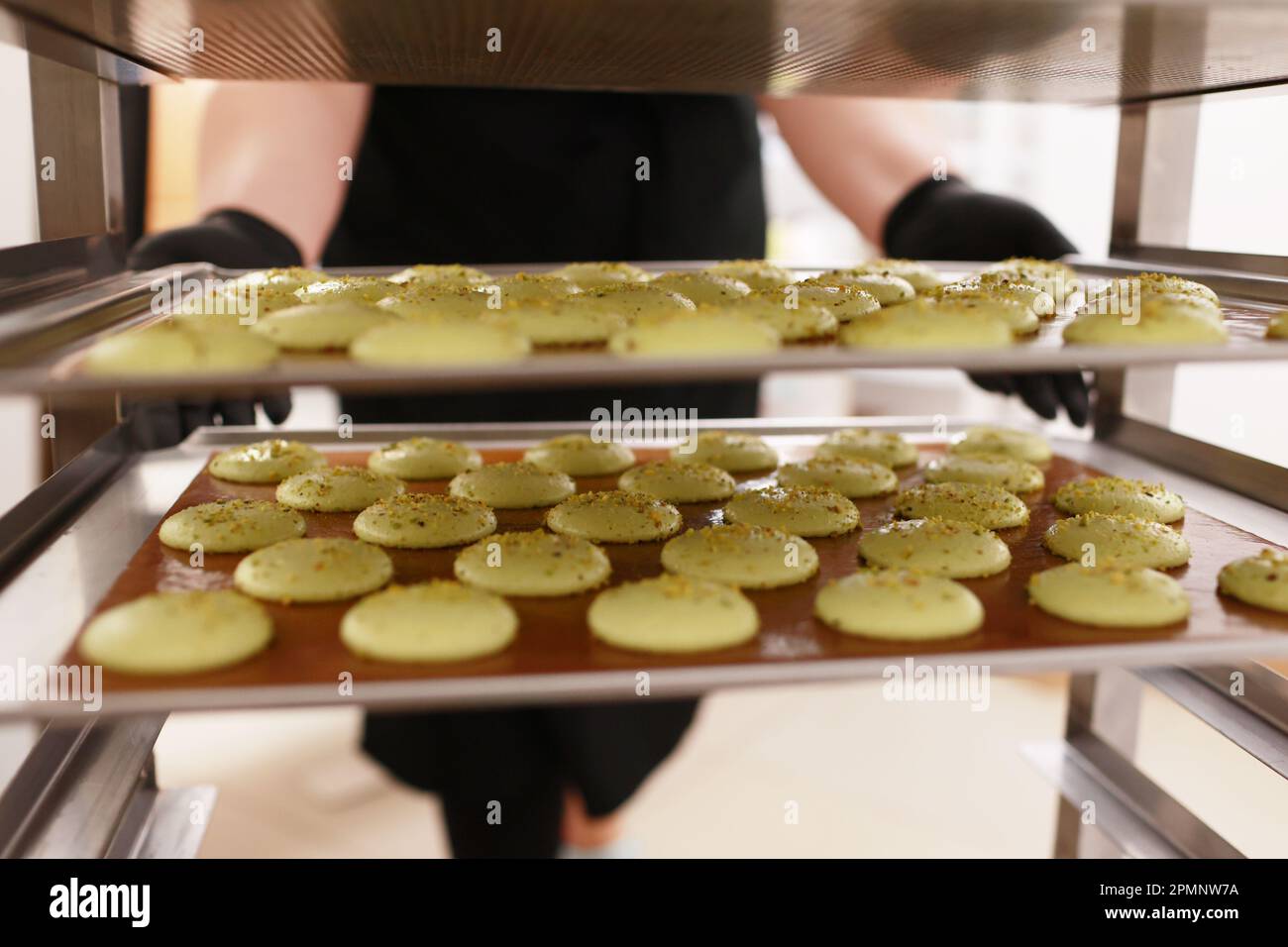 Der Koch stellt ein Tablett voller bunter Makronen auf ein Metallregal mit weiteren Makronen, die sich in einer Bäckerei befinden. Zubereitung köstlicher französischer Desser Stockfoto