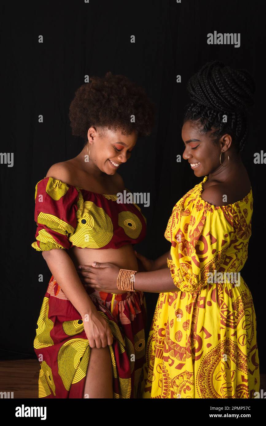 Zwei Freundinnen, eine davon schwanger, zusammen in bunten Kleidern. Isoliert auf gelbem Hintergrund. Stockfoto