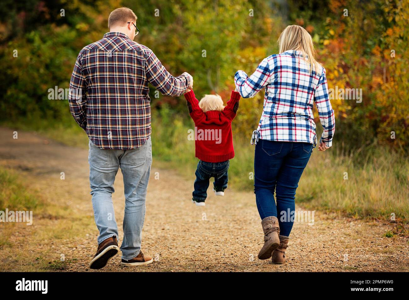 Mutter und Vater halten mit dem kleinen Sohn Händchen, während sie im Herbst in einem Stadtpark in Edmonton, Alberta, Kanada, einen Pfad entlang gehen Stockfoto