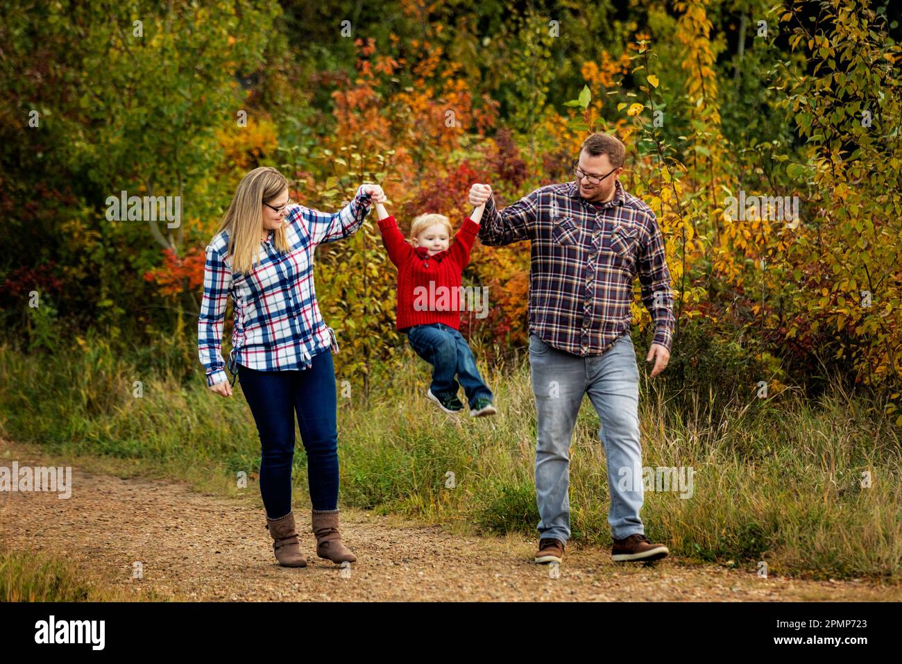 Mutter und Vater halten mit dem kleinen Sohn Händchen, während sie im Herbst in einem Stadtpark in Edmonton, Alberta, Kanada, einen Pfad entlang gehen Stockfoto