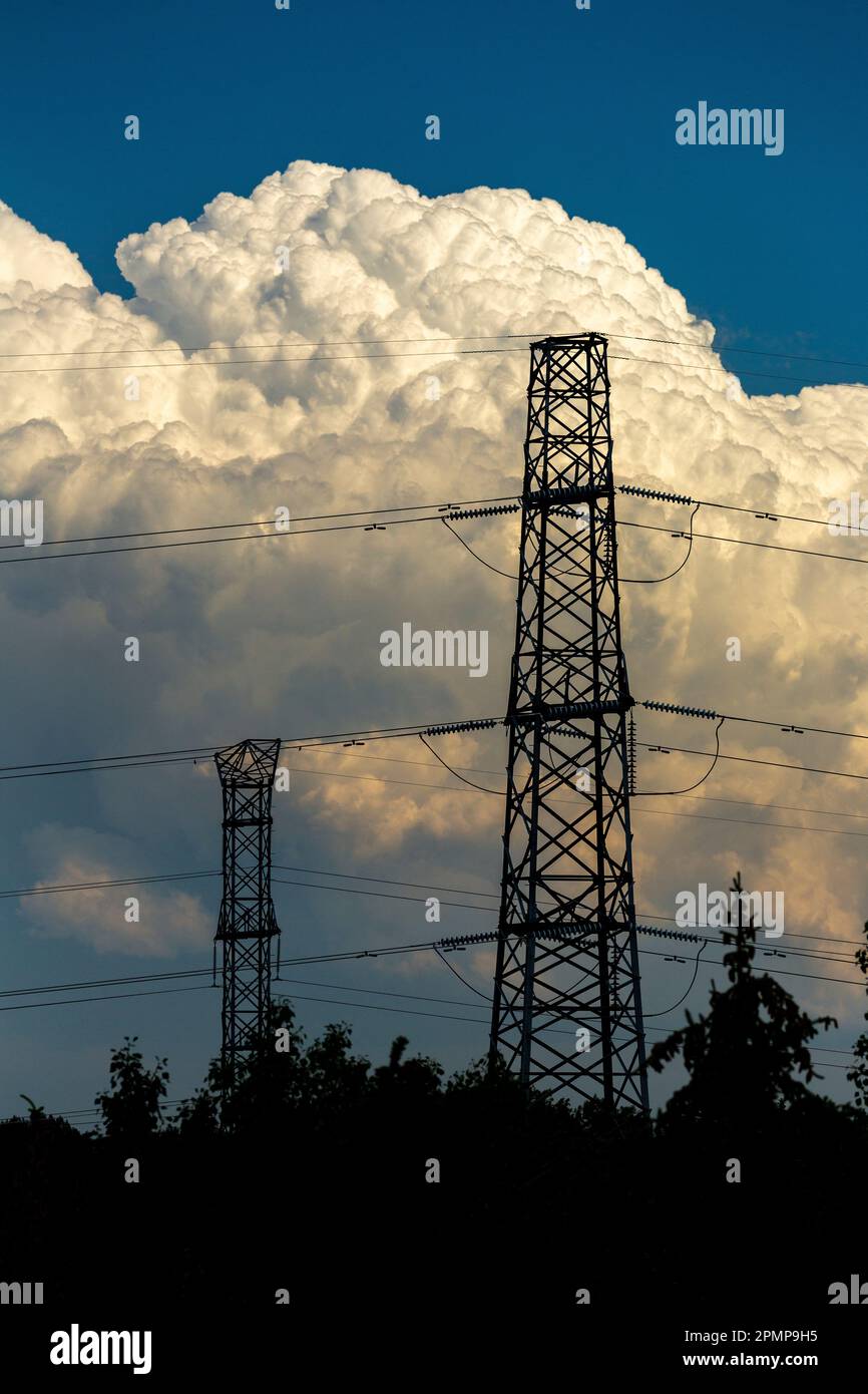 Silhouette aus elektrischen Metalltürmen mit Bäumen im Vordergrund und dramatischen Sturmwolken im Hintergrund und blauem Himmel darüber Stockfoto