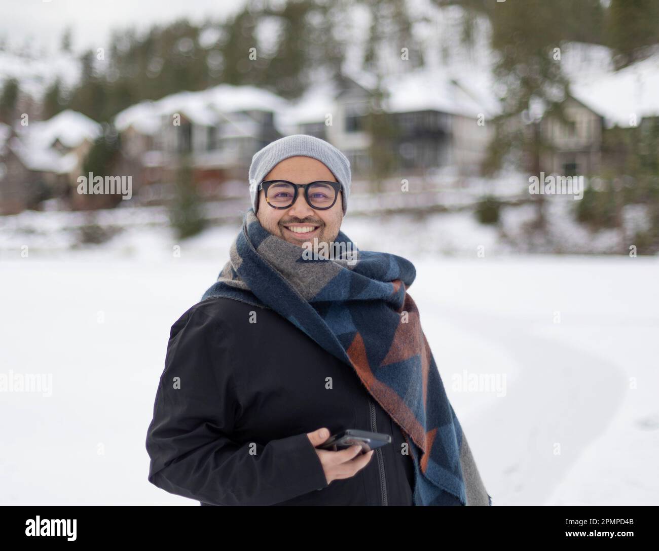 Außenporträt eines jungen Mannes an einem Wintertag, der eine Brille, einen Mantel und einen Strickmütze trägt und ein Smartphone in der Hand hält; British Columbia, Kanada Stockfoto