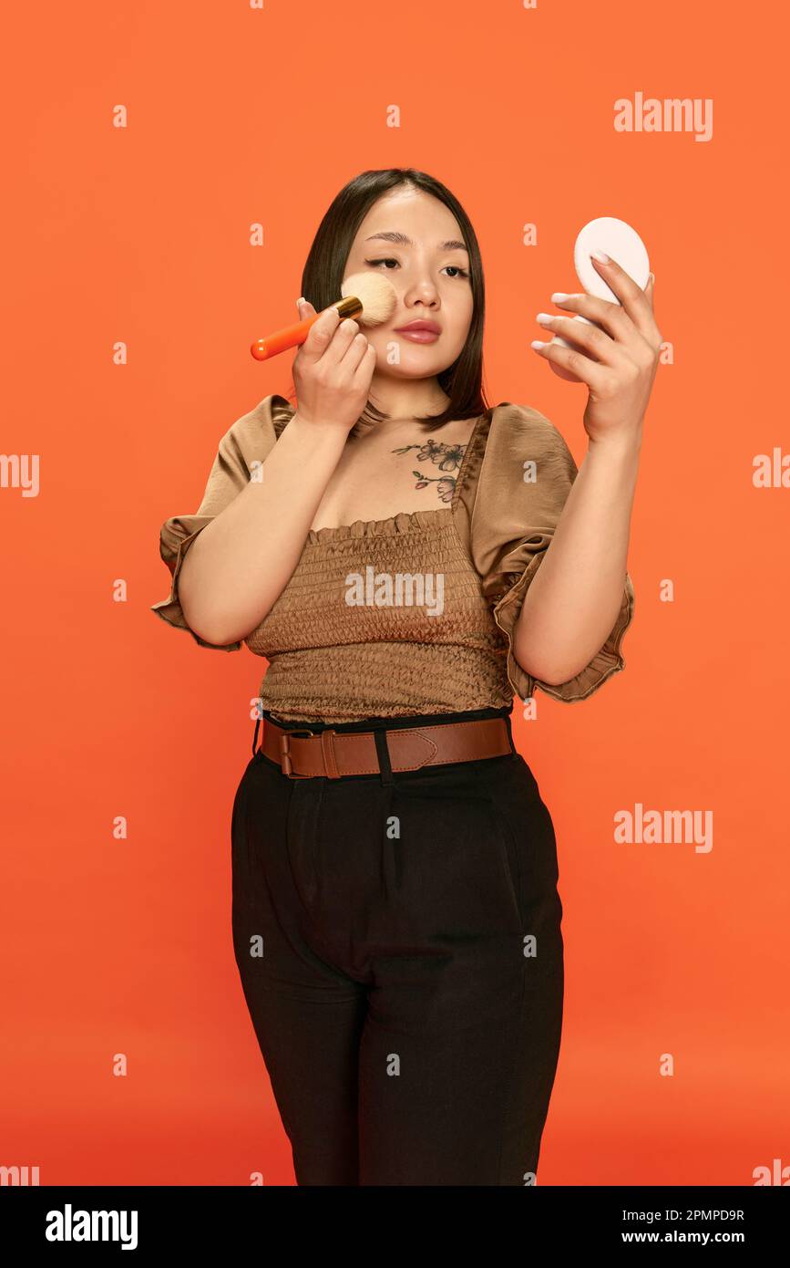 Porträt eines jungen, wunderschönen asiatischen Mädchens in stilvollen Kleidern, das sich schminkt und vor orangefarbenem Studiohintergrund Rouge auf die Wangen trägt Stockfoto