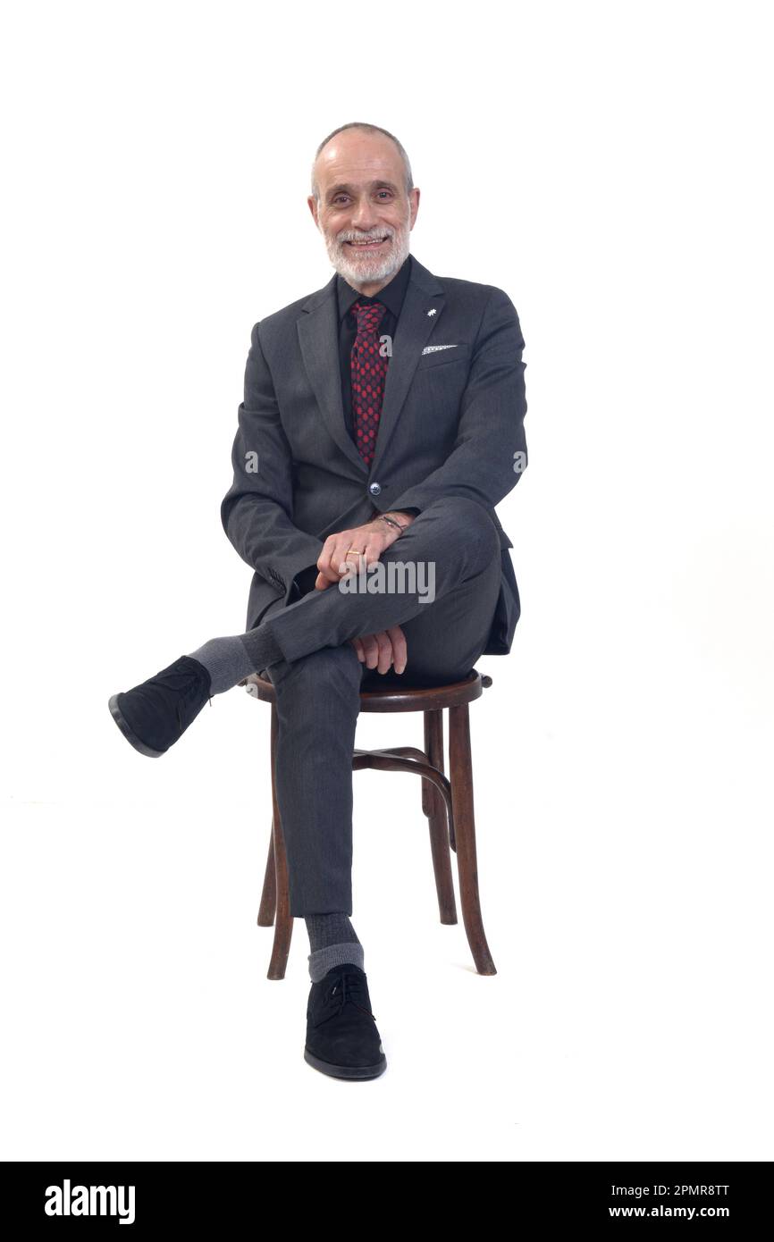 Vorderansicht eines Mannes, der auf einem Stuhl sitzt, mit Anzug und Krawatte und gekreuzten Beinen lächelt und auf die Kamera auf weißem Hintergrund schaut Stockfoto