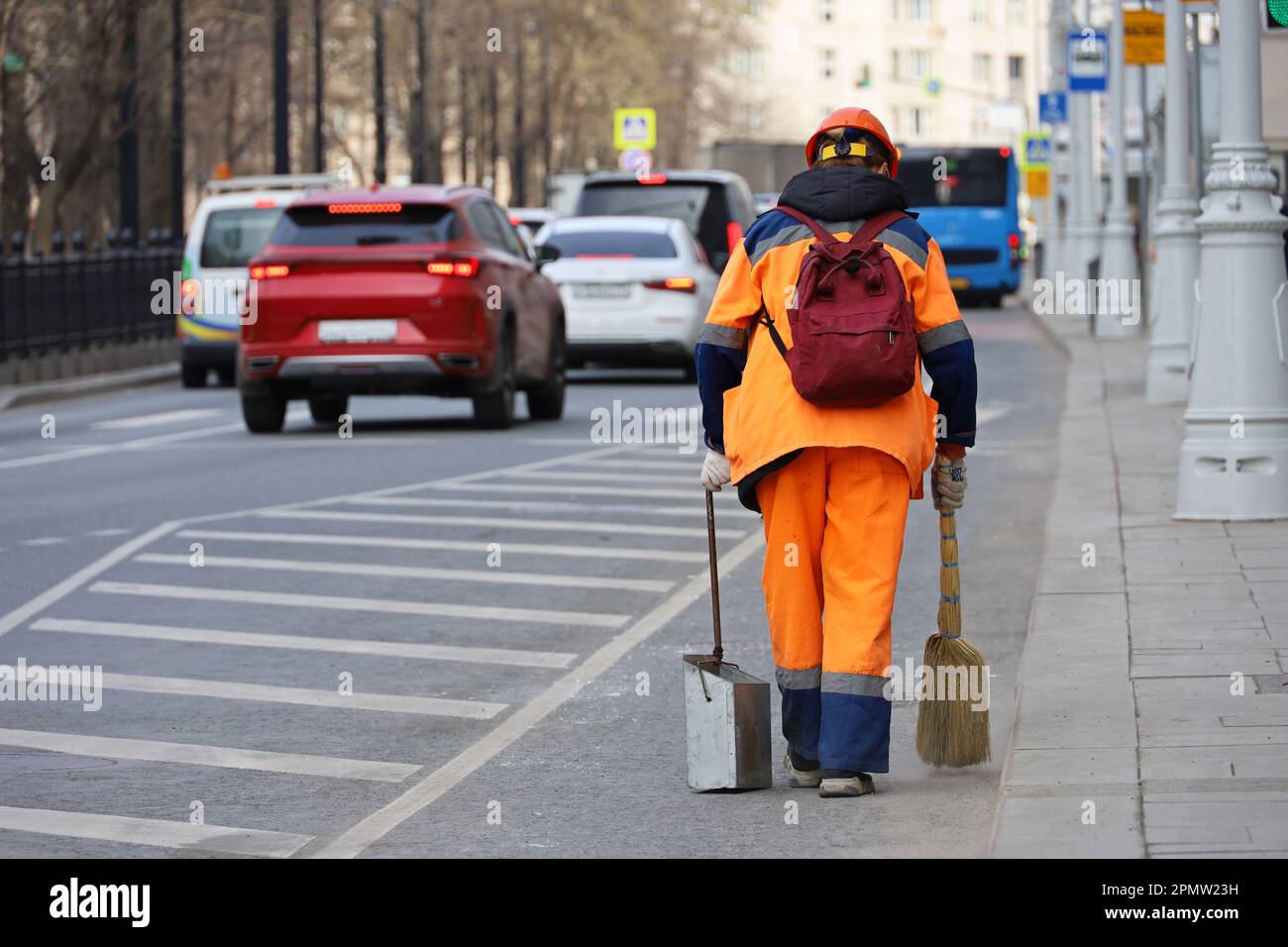 Eine Arbeiterin in orangefarbener Uniform mit Besen fegt die Straße auf dem Hintergrund von Autos. Straßenreinigung in der Frühjahrsstadt Stockfoto