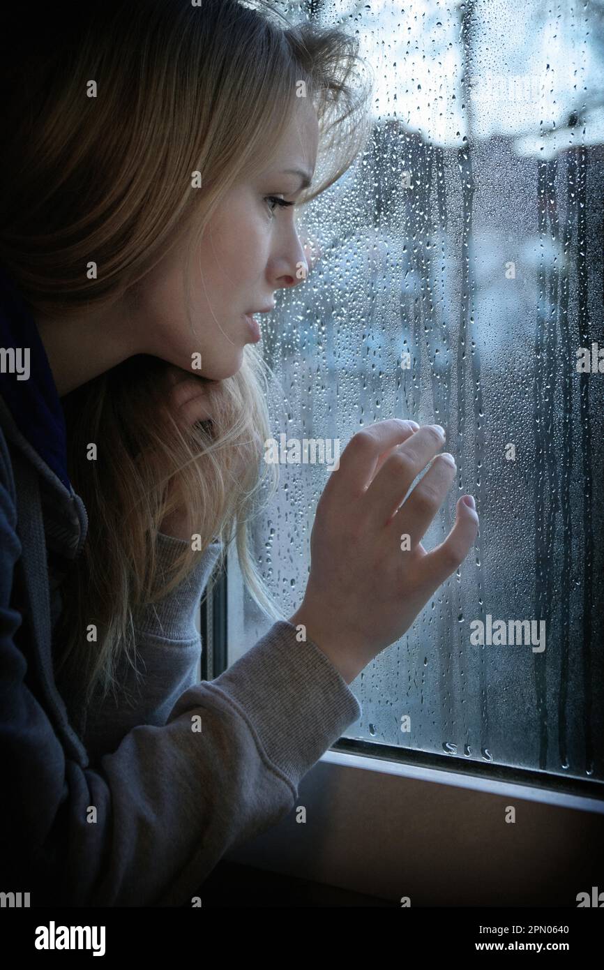 Melancholische junge Frau, die durch ein Fenster blickt, mit Regentropfen und zusätzlichem Filterkorn und Vignette Stockfoto