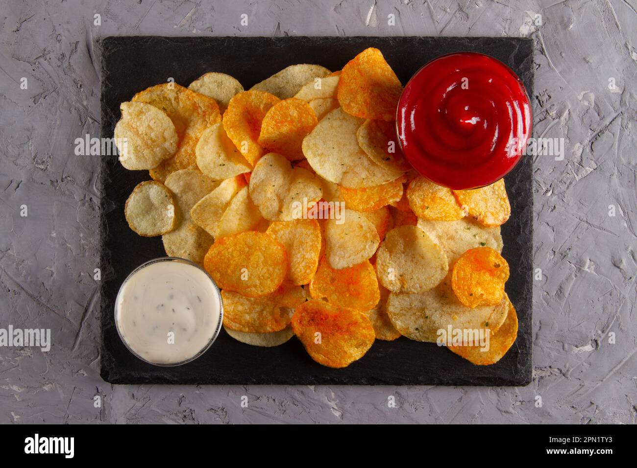 Ansicht von oben auf den Haufen Kartoffelchips mit zwei Dippsaucen auf schwarzem Schiefer auf einem grauen Gipstisch. Klassische Partysnacks Stockfoto