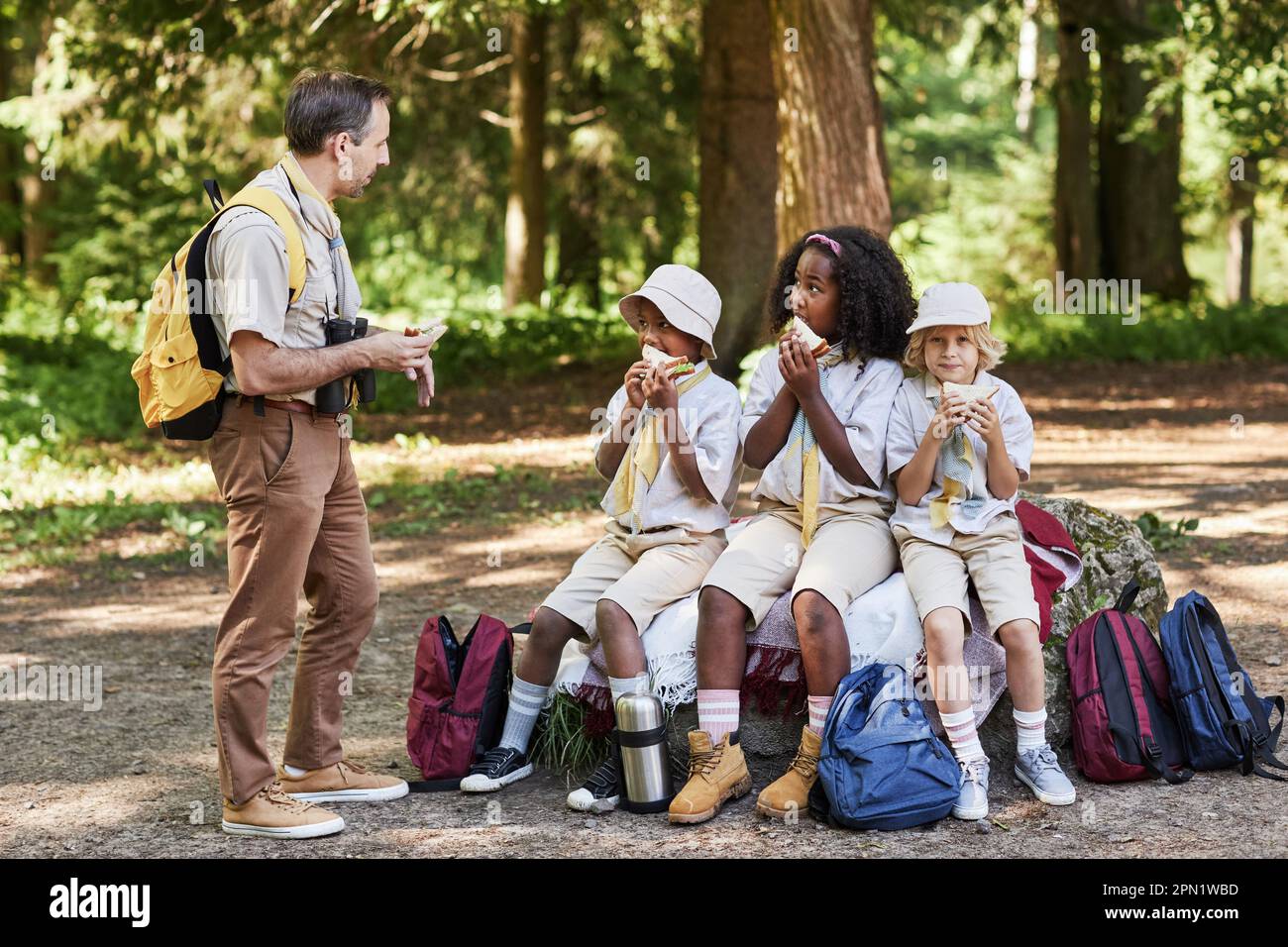 Vielfältige Gruppe von Spähern, die während der Wanderung eine Mittagspause genießen, während ein erwachsener Führer mit Kindern spricht Stockfoto