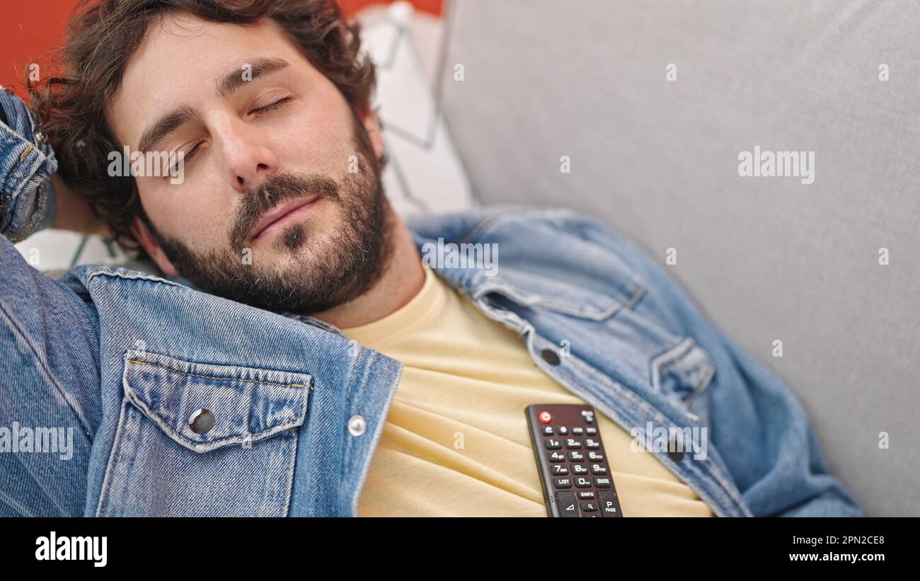 Junger hispanischer Mann mit tv-Fernbedienung, der auf dem Sofa liegt und zu Hause schläft Stockfoto