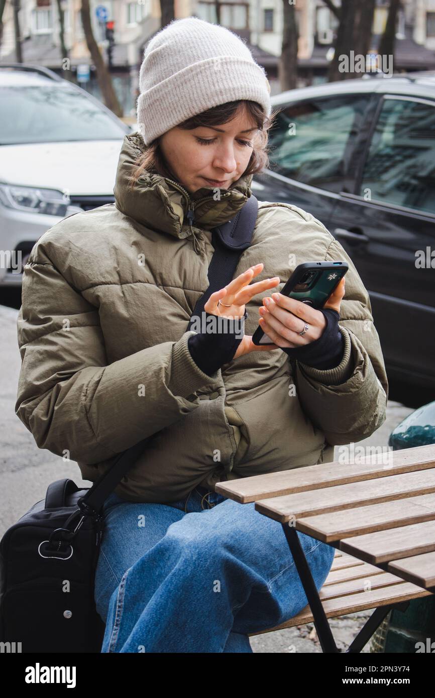 Ein Mädchen, das im Café auf dem Bürgersteig telefoniert. Frau in warmen Kleidern sitzt im Straßencafé mit Telefon. Großstadtleben. Moderne Technik im Alltag Stockfoto