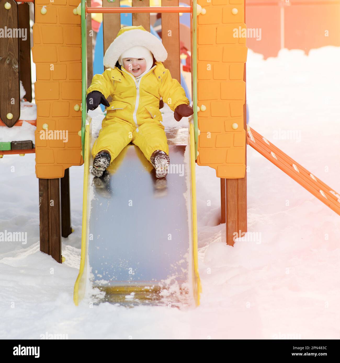 Der kleine Junge fährt auf einer Rutsche und spielt auf einem Winterspielplatz. Ein Kind in einem gelben Overall auf einer Kinderrutsche im Schnee. Ein Kind im Alter von einem Jahr Stockfoto