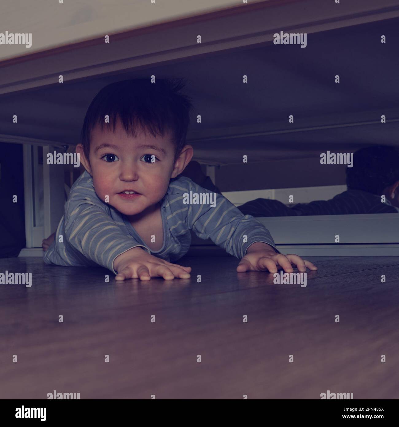 Das Baby krabbelt und versteckt sich unter dem Bett. Das Kind kletterte unter das Sofa und setzte sich auf den Boden. Ein Junge im Alter von einem Jahr, acht Monate, volle Länge Stockfoto